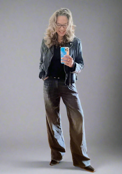A person with long hair and glasses captures a mirror selfie. They are clad in a black leather jacket and dark shirt, complemented by the R13 Damon Pleated Jeans featuring a vintage wash. Holding a phone encased in vibrant colors, they pose against a plain backdrop.