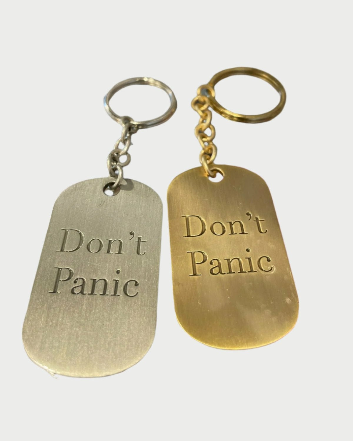 Two keychain dog tags are displayed on a glass surface. Both are rectangular with rounded edges, resembling vintage designs. One is silver brass with the inscription "Don't Panic" in black, and the other is gold with the same inscription in green. These are part of the Don’t Panic key chain collection by Simply Eartha.
