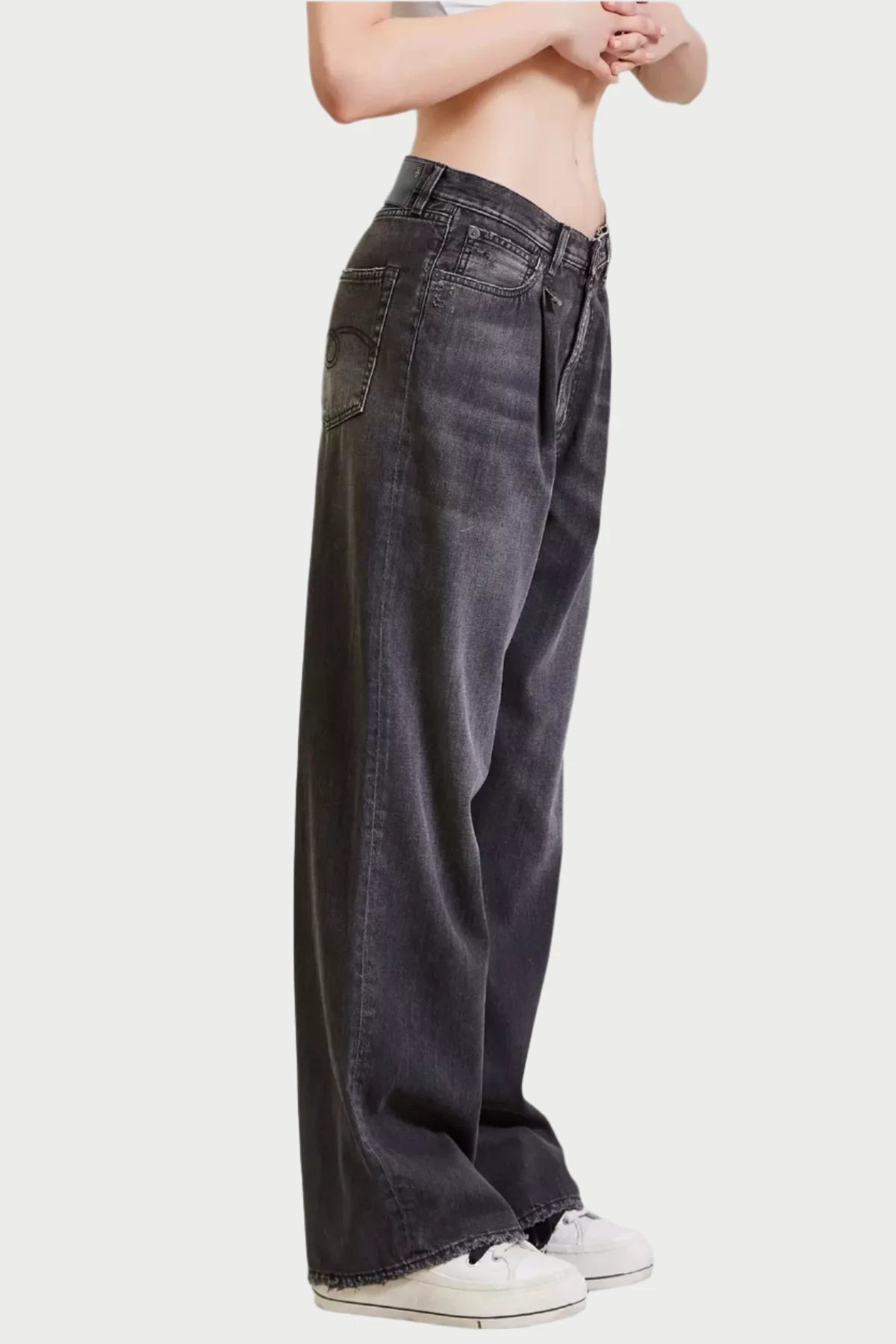 A person stands with folded hands, wearing R13 Damon Pleated Jeans in a vintage wash and white sneakers. The plain background emphasizes the casual outfit.