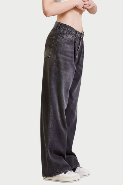 A person stands with folded hands, wearing R13 Damon Pleated Jeans in a vintage wash and white sneakers. The plain background emphasizes the casual outfit.